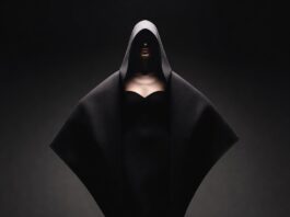 Model in dramatic oversized cocooning coat on a pitch-black runway with chiaroscuro spotlight lighting representing the Balenciaga runway 2026 ClairObscur collection by Pierpaolo Piccioli at Paris Fashion Week