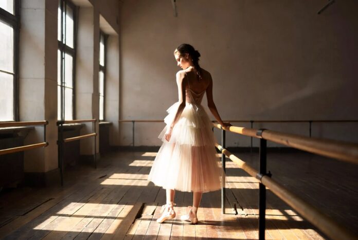 ballet-luxury-fashion-ballerina-runway-trend-2026 Model in a layered blush tulle gown and ribbon-tied ballet flats posed at a barre in a sunlit ballet studio, illustrating the ballet luxury fashion trend dominating 2026 couture and runway collections.