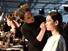 Makeup artist applying editorial runway beauty look to a model backstage at a fashion week show, representing the Beauty Trends Hub covering runway makeup, hair, and skincare trends