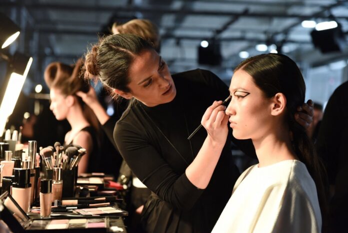 beauty-trends-hub-runway-makeup-hair-skincare-fashion-week Makeup artist applying editorial runway beauty look to a model backstage at a fashion week show, representing the Beauty Trends Hub covering runway makeup, hair, and skincare trends