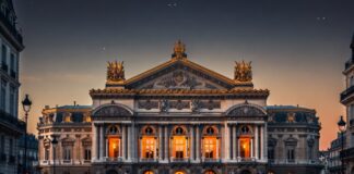 Paris Opera Ballet’s Radical New Season Is Dividing the Dance World The Palais Garnier opera house in Paris at dusk, golden interior light through arched windows, the ornate neoclassical facade representing the Paris Opera Ballet's 2026 season at the center of a global classical versus contemporary ballet debate.