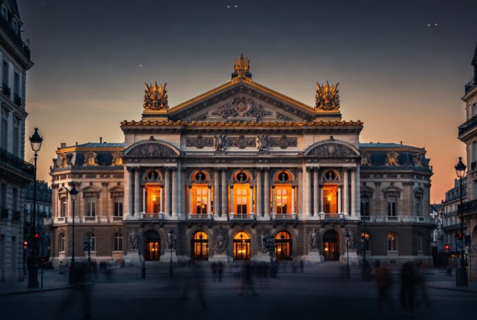 Paris Opera Ballet 2026 — Palais Garnier at Dusk The Palais Garnier opera house in Paris at dusk, golden interior light through arched windows, the ornate neoclassical facade representing the Paris Opera Ballet's 2026 season at the center of a global classical versus contemporary ballet debate.