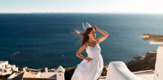 European Coastal Style Is Influencing Every Vacation Outfit This Summer Woman in a white linen dress standing on whitewashed Mediterranean steps with blue sea views.
