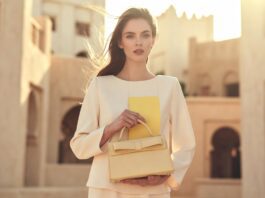 Dior Cruise 2026 Is Taking Over Summer With Elegant Glamour A model in soft cream resort tailoring holding a structured top-handle bag in warm summer light