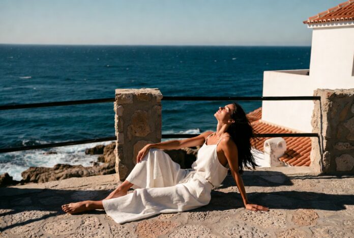 Woman in a white wide-leg linen set on a sun-drenched European terrace overlooking a blue sea.