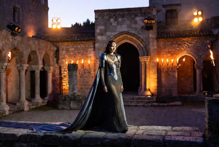 Louis Vuitton Cruise 2026 Avignon Female Knight Show Models in armor-inspired metallic gowns on a stone runway inside a gothic medieval palace courtyard at dusk