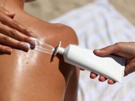 Sunscreen being applied to bare shoulders in bright summer sunlight, with a tube of SPF visible beside.