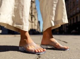 Jelly Shoes, Beaded Sandals and ’90s Footwear Are Fueling Summer Street Style Woman wearing translucent jelly sandals with a cream linen co-ord on a sunlit summer city street.
