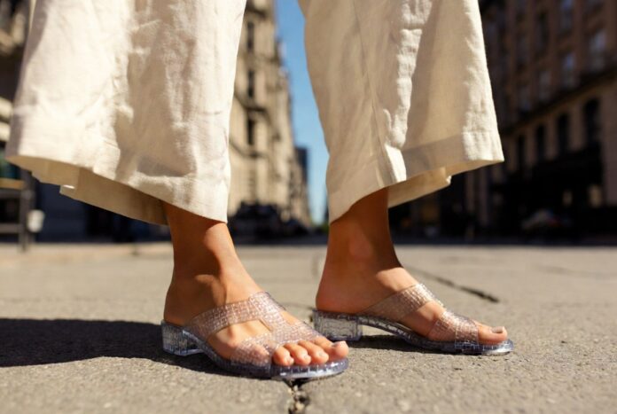 Woman wearing translucent jelly sandals with a cream linen co-ord on a sunlit summer city street.