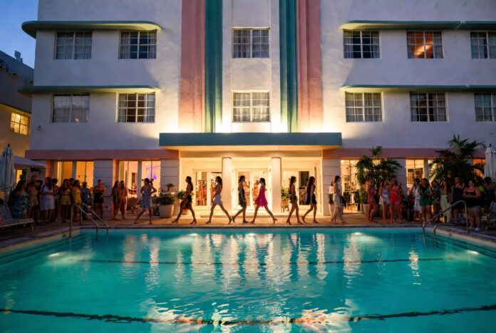 Models on a runway built over an Art Deco hotel pool in warm evening light representing PARAISO RISE Miami Swim Week 2026 at the Kimpton Surfcomber Hotel and the immersive fashion experience format redefining pool runway fashion shows