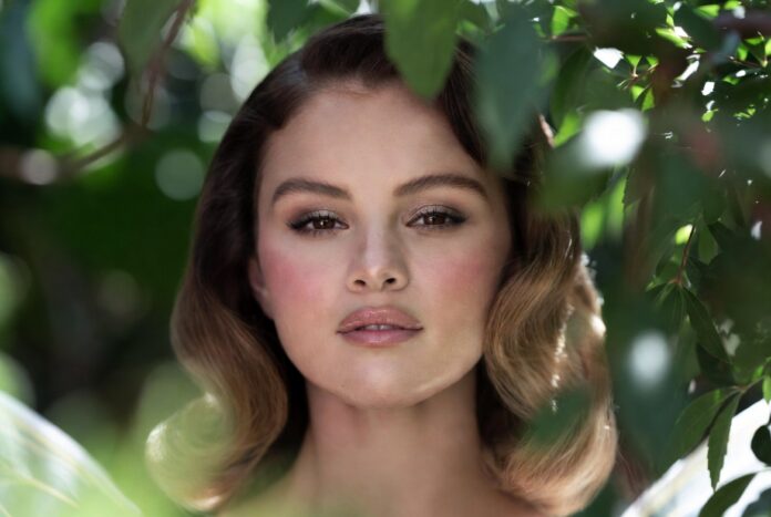 Woman in white tulle dress in a lush garden with rosy pink blush shimmering eyeshadow ultra-glossy lips and soft waves representing the Selena Gomez Rare Beauty fairy makeup aesthetic and garden photoshoot beauty look from the June 2026 campaign