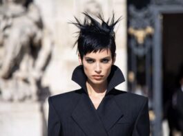 A woman with a dramatic dark rough-cut pixie hairstyle in a tailored suit outside a couture show